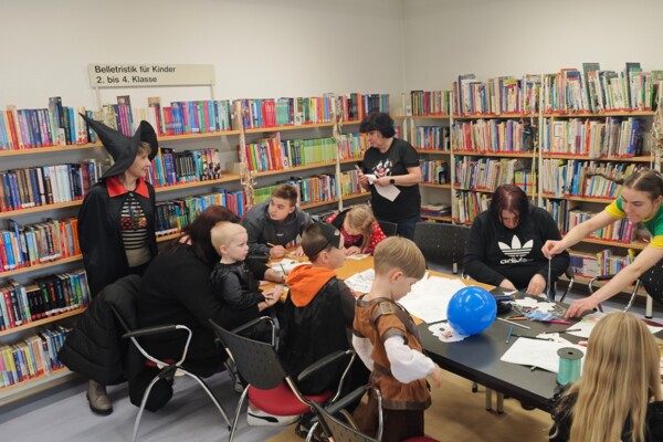 Faschingsbasteln 2026 in der Bibliothek Blick in die Kinderabteilung der Schmöllner Bibliothek beim Faschingsbasteln über eine lange Tafel an der bastelnde Kinder und Eltern sitzen und die Mitarbeiterinnen der Bibliothek um den Tisch herum tatkräftig unterstützen