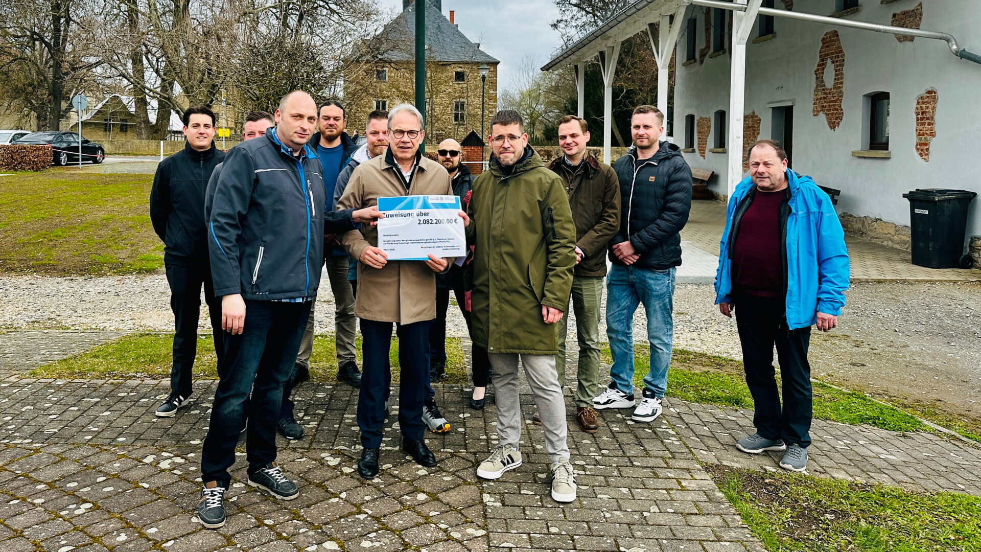 Gruppenbild bei der Übergabe der Eingliederungsprämie in Dobitschen mit dem Ortsteilbürgermeister Björn Steinicke, Thüringens Innenminister Georg Maier, Schmöllns Bürgermeister Sven Schrade in der ersten Reihe sowie Kolleg*innen aus der Stadtverwaltung, dem Stadtrat und dem Ortsteilrat im Hintergrund auf dem Dorfplatz