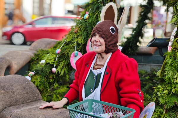 Blick auf die Osterhäsin Astrid Pohl, die im Brunnen vor der geschmückten Ostergirlande kniet, sich mit ihren Händen auf den Rand stützt und jemanden anlächelt 