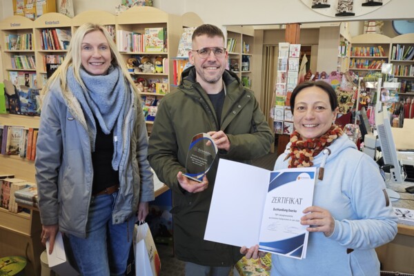 Platz 1 Top Akzeptanzstelle: Buchhandlung Goerke Julia Waldmann, Wirtschaftsförderin, Bürgermeister Sven Schrade, der einen kleinen Pokal hochhält, und Buchhändlerin Kristin Mielke, die ein Zertifikat hochhält, stehen im Eingangsbereich der Buchhandlung Goerke in Schmölln