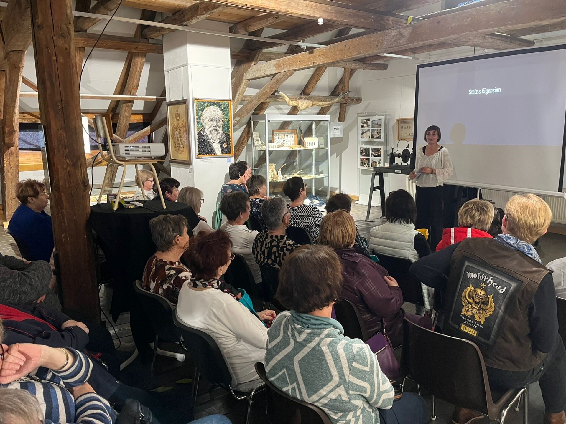 Blick in einen Ausstellungsraum über die Köpfe der Besucher*innen der Filmvorführung "Stolz und Eigensinn" im Schmöllner Knopf-und Regionalmuseum zum internationalen Frauentag 2026 auf eine weiße Leinwand, vor der die neue Museumsleiterin Constance Böhme steht und eine Rede hält