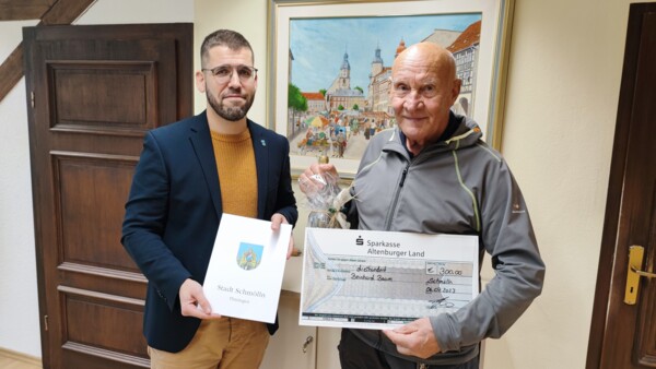 Bürgermeister Sven Schrad steht neben Bernhard Baum. Sven Schrade hält die Urkunde für Herrn Baum in der Hand. Herr Baum hält den Scheck in der Hand.