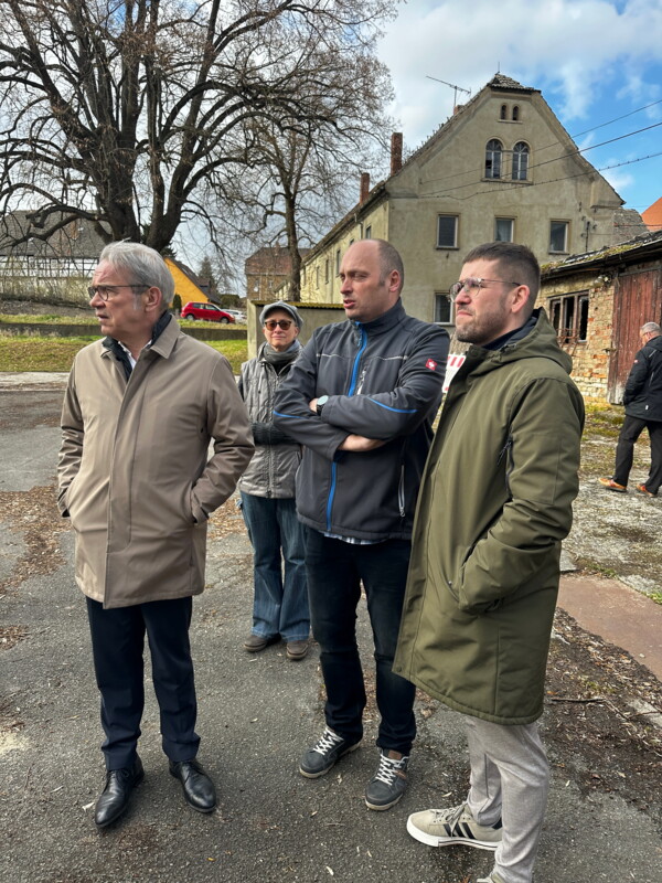 Blick auf den Thüringer Innenminister Georg Maier, den Ortsteilbürgermeister Björn Steinicke und den Schmöllner Bürgermeister Sven Schrade, die auf dem Dobitschner Dorfplatz zusammenstehen, sich unterhalten und sich umsehen