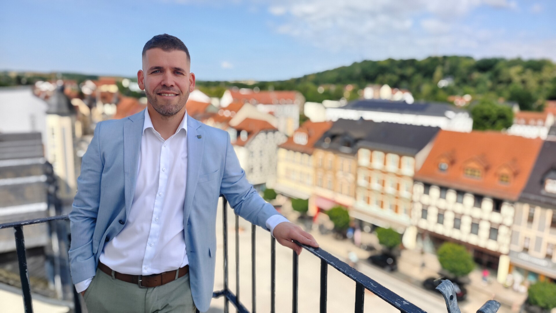 Bild des Schmöllner Bürgermeisters Sven Schrade mit einem weißen Hemd, einer lilanen Krawatte und einem schwarzen Jackett auf den Stufen zum Haupteingang des Schmöllner Rathauses