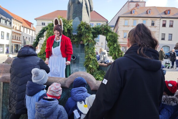 Blick über eine Kindergartengruppe mit den Erzieher*innen auf Osterhäsin Astrid Pohl, die im Schmöllner Marktbrunnen steht und sich lächelnd mit einer Erzieherin unterhält 