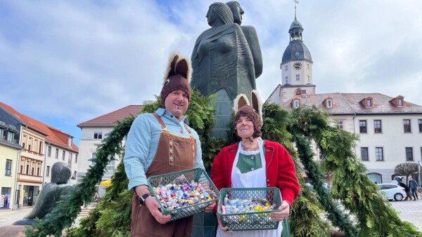Die beiden Osterhasen, Stefan Klein und Astrid Pohl, stehen mit vollen Süßigkeitsbehältern im Schmöllner Marktbrunnen, der mit der grünen Ostergirlande geschmückt ist