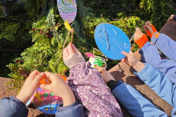 Blick von oben auf mehrere Hände der Kindergartenkinder, die ihre selbstgebastelten Osterdekorationen für den Marktbrunnen, darunter viele bunte Ostereier, über den Rand des Brunnens hinhalten 