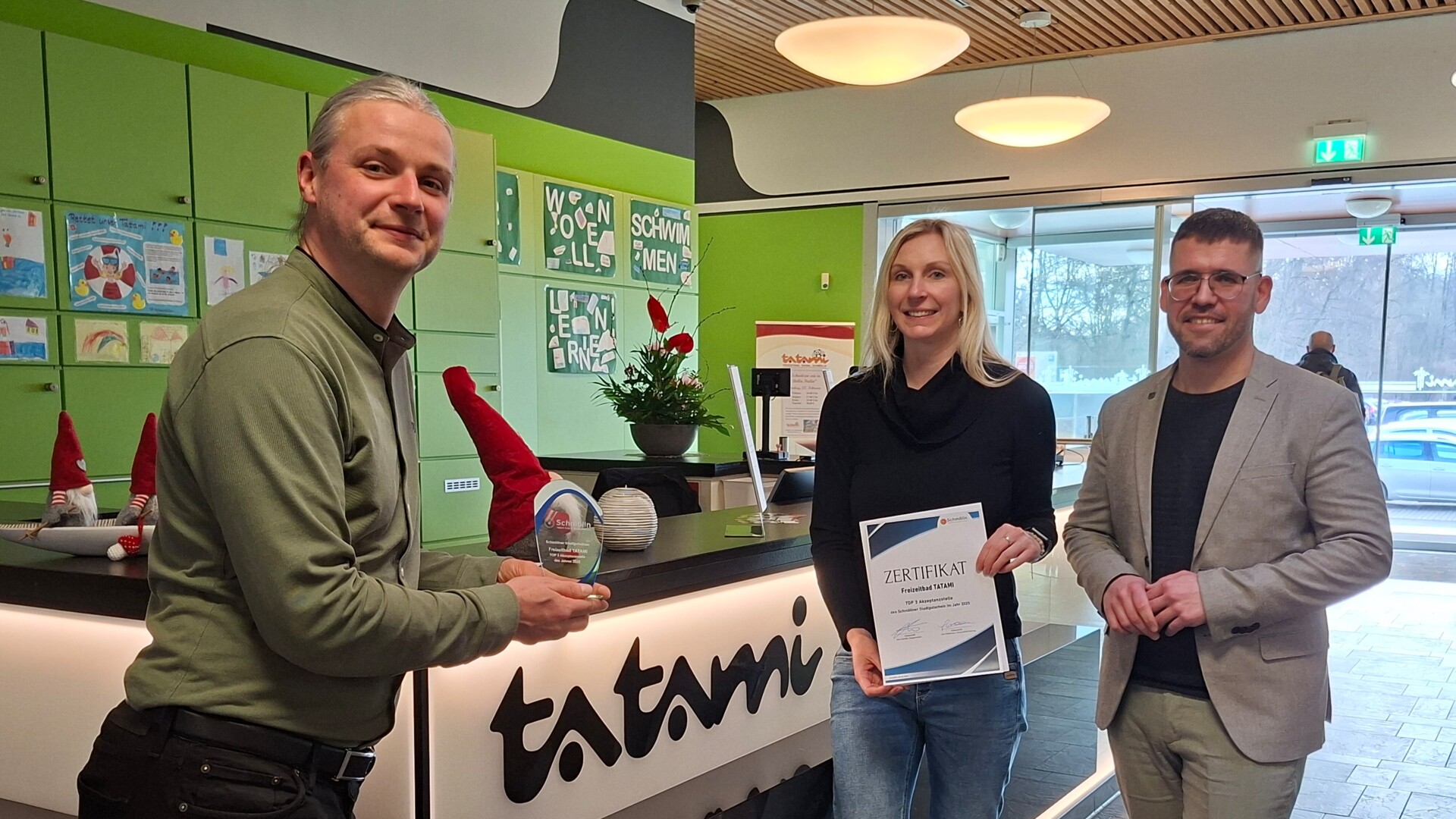 Platz 3 Top Akzeptanzstellen: Freizeitbad TATAMI Severin Kühnast, Geschäftsführer der Schmöllner Stadtwerke, der einen kleinen Pokal hochhält, Julia Waldmann, Wirtschaftsförderin, die ein Zertifikat hochhält, und Bürgermeister Sven Schrade stehen an der Rezeption im Eingangsbereich des Tatami