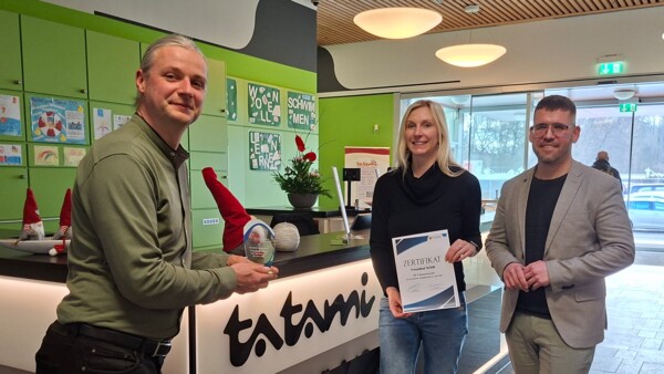 Severin Kühnast, Geschäftsführer der Schmöllner Stadtwerke, der einen kleinen Pokal hochhält, Julia Waldmann, Wirtschaftsförderin, die ein Zertifikat hochhält, und Bürgermeister Sven Schrade stehen an der Rezeption im Eingangsbereich des Tatami