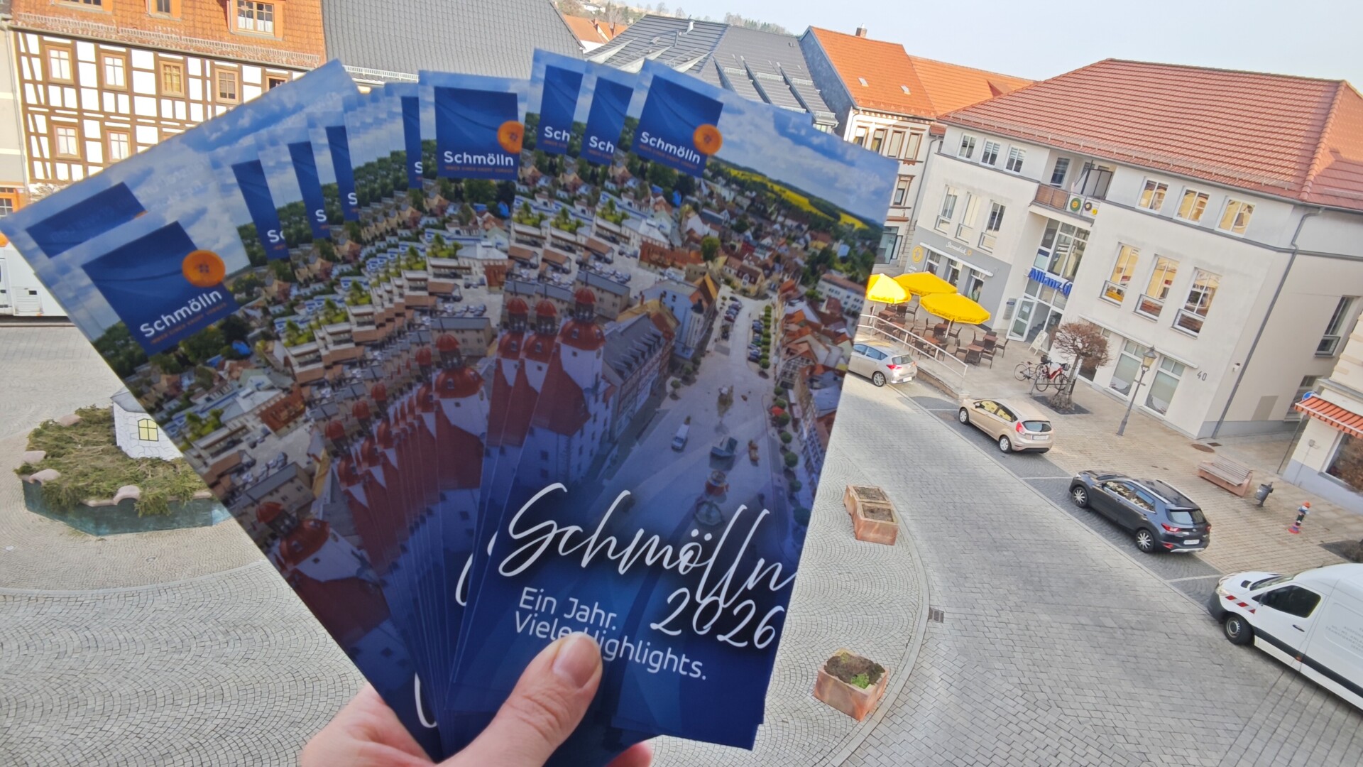 Blick auf mehrere aufgefächerte Veranstaltungsflyer für Schmölln 2026, die den Schmöllner Markt aus der Vogelperspektive zeigen mit Schriftzug "Schmölln 2026 - Ein Jahr. Viele Highlights.", die aus einem Fenster des Schmöllner Rathauses, das auf den Markt hinausgeht, rausgehalten werden