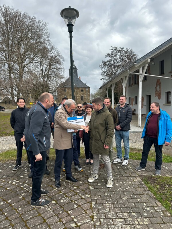Gruppenbild bei der Übergabe der Eingliederungsprämie in Dobitschen mit dem Ortsteilbürgermeister Björn Steinicke und Thüringens Innenminister Georg Maier, der dem Schmöllner Bürgermeister Sven Schrade in der ersten Reihe den Scheck überreicht und die Hand schüttelt, mit Kolleg*innen aus der Stadtverwaltung, dem Stadtrat und dem Ortsteilrat im Hintergrund auf dem Dorfplatz
