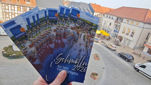 Blick auf mehrere aufgefächerte Veranstaltungsflyer für Schmölln 2026, die den Schmöllner Markt aus der Vogelperspektive zeigen mit Schriftzug "Schmölln 2026 - Ein Jahr. Viele Highlights.", die aus einem Fenster des Schmöllner Rathauses, das auf den Markt hinausgeht, rausgehalten werden